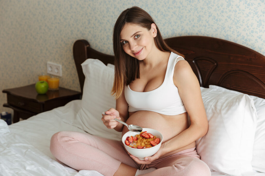 Portrait of a smiling young pregnant woman having healthy breakfast while sitting on bed with a bowl and spoon