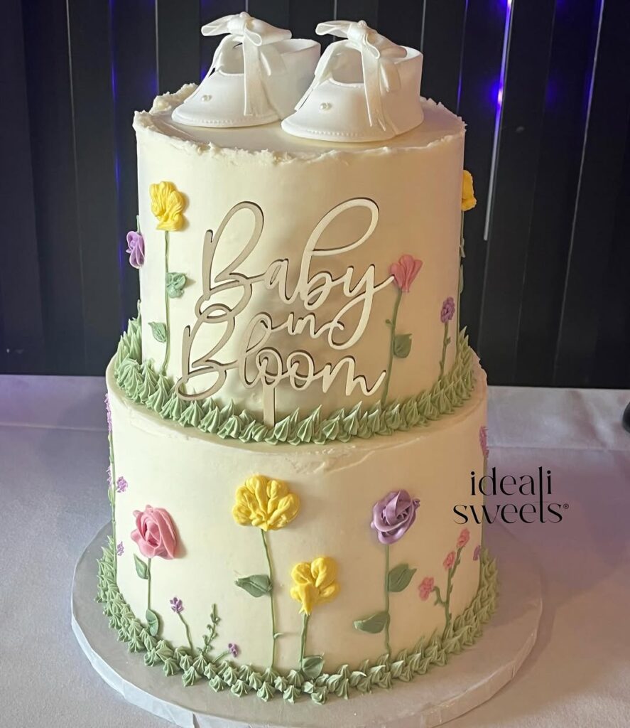 A two-tier cake decorated with flowers, green frosting, and white baby shoes. Text says "Baby in Bloom."