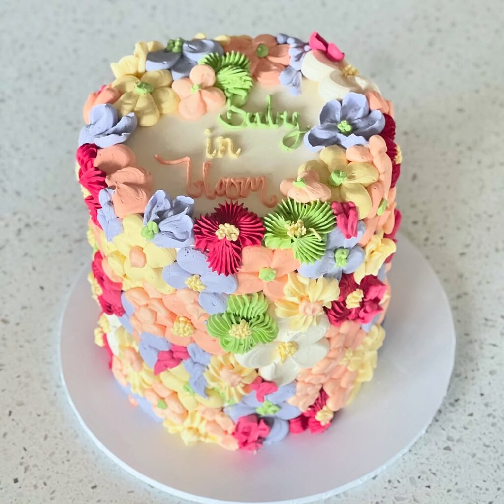 A colorful cake decorated with frosting flowers in pink, yellow, and purple, with the words 'Baby in Bloom' on top.