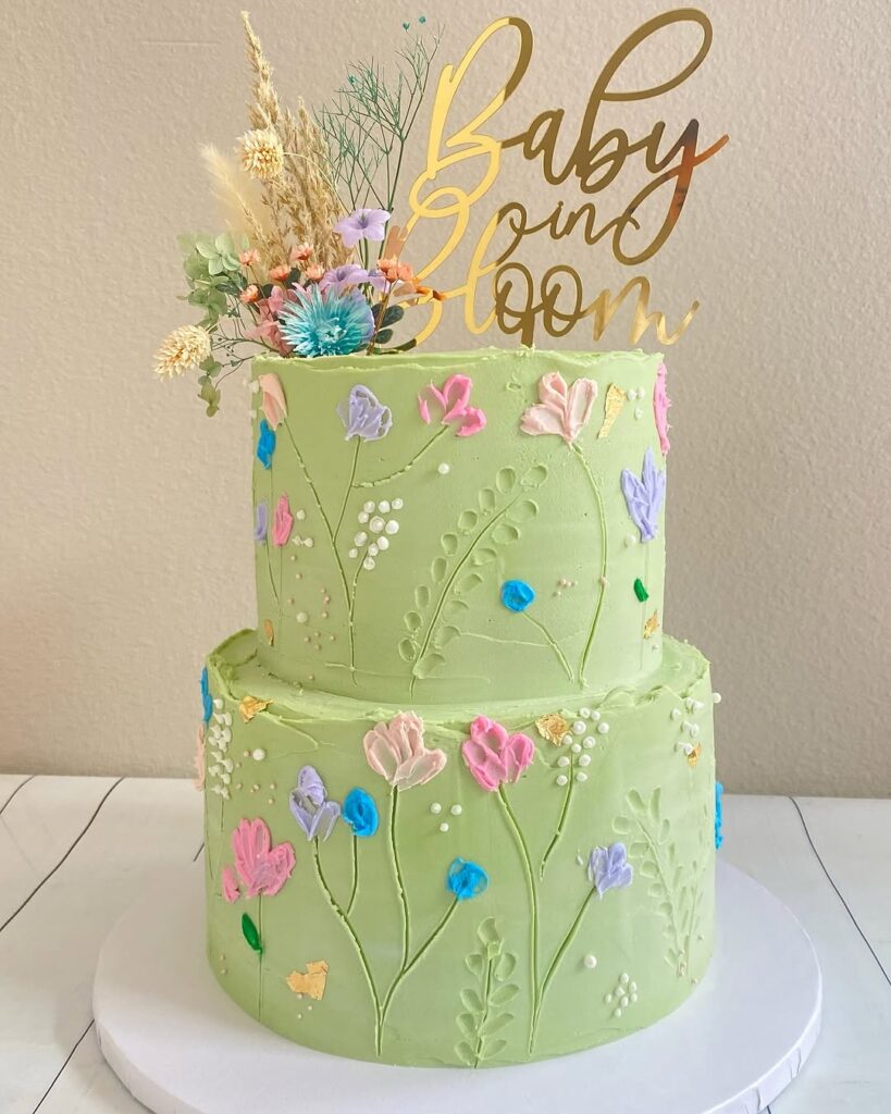 A two-tier green cake decorated with colorful flowers and a gold 'Baby in Bloom' topper.