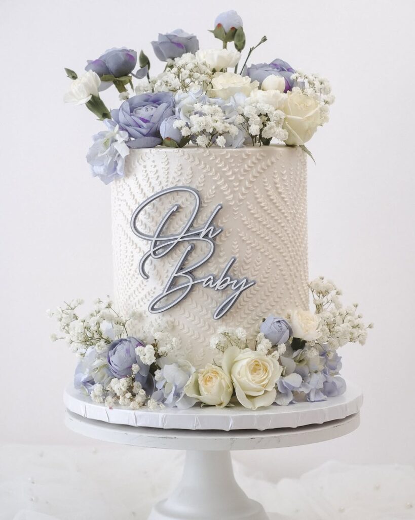 A tall cake decorated with blue flowers and white roses, with the text 'Oh Baby' on it.