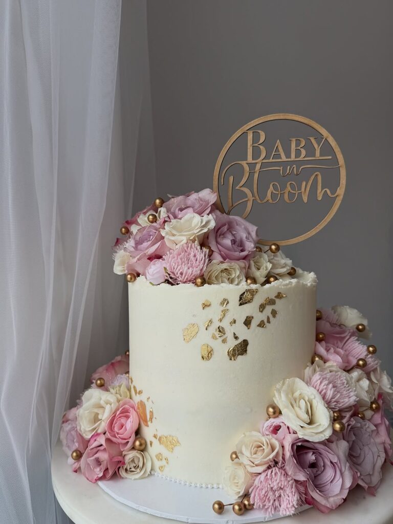 A tall white cake decorated with pink flowers and gold pearls, with a sign that says Baby in Bloom