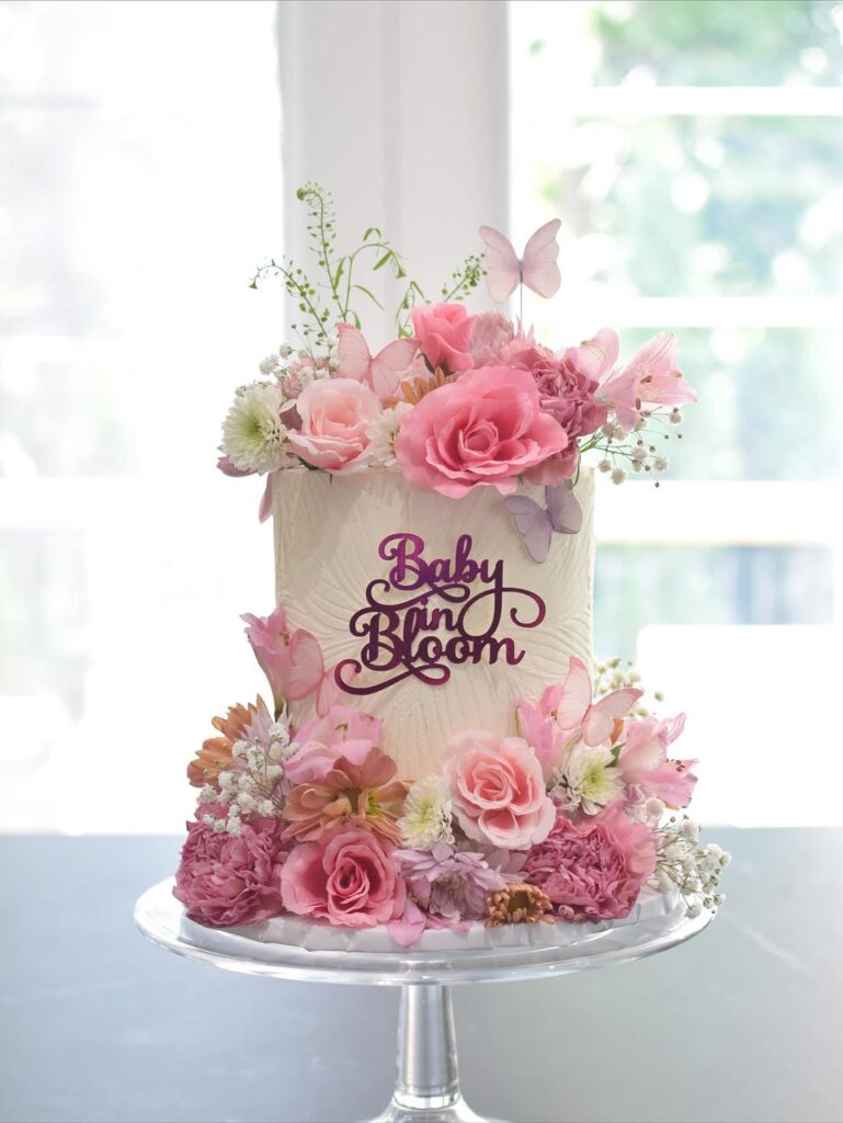 A tall white cake decorated with pink flowers and butterflies, with a sign that says Baby in Bloom