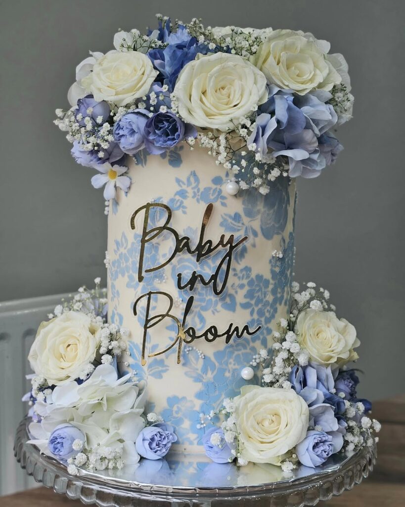 A tall cake decorated with blue flowers and white roses, with the text 'Baby in Bloom' on it.