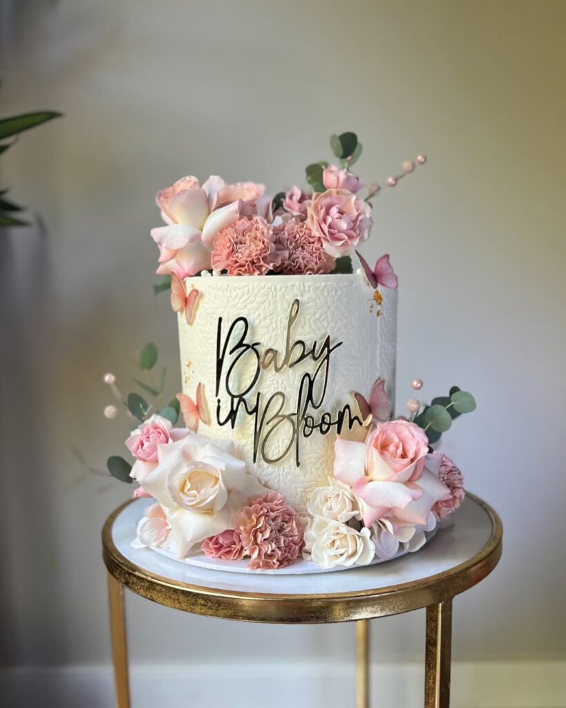 A tall white cake decorated with pink flowers, with a sign that says Baby in Bloom
