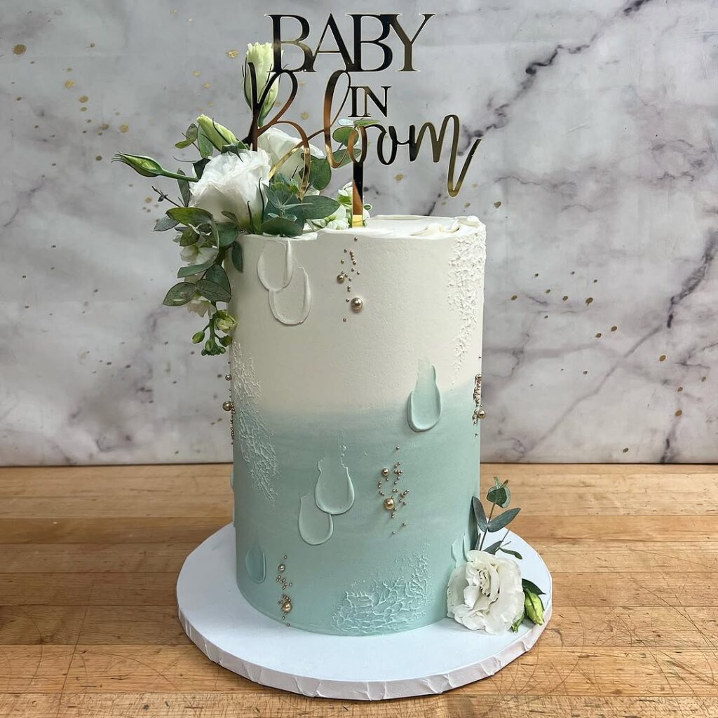 A tall cake in blue and white with floral decorations and a gold 'Baby in Bloom' topper.