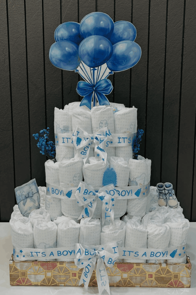 three-tiered diaper cake, featuring blue and white "It's a Boy!" ribbons and adorable balloon toppers